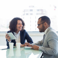 Joyful successful mentor training intern. Happy business woman showing tablet screen to male colleague, he writing notes in notebook.