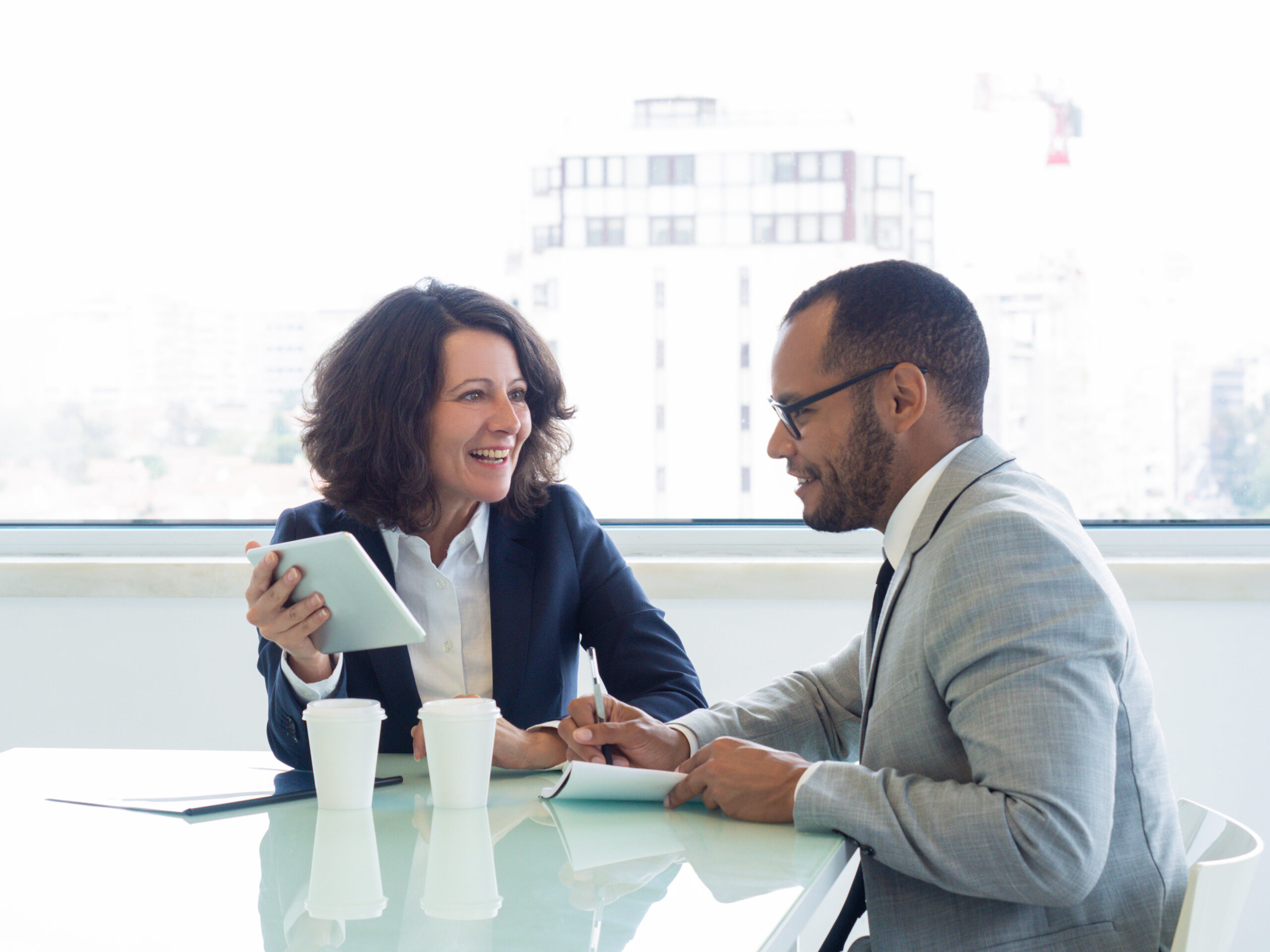 Joyful successful mentor training intern. Happy business woman showing tablet screen to male colleague, he writing notes in notebook.